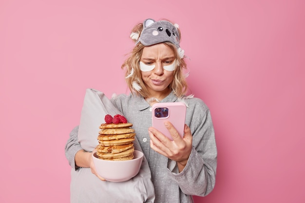 Serious beautiful woman awakes early in morning looks attentively at smartphone screen checks newsfeed wears blindfold and pajama holds bowl of tasty pancakes applies beauty patches under eyes