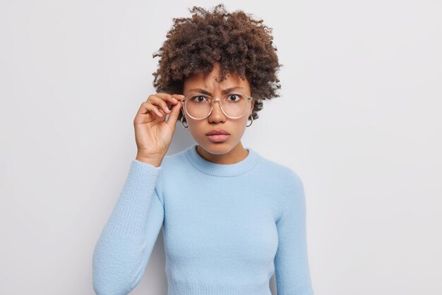 Serious beautiful woman with dark curly hair looks attentively at camera through transparent glasses dressed in casual blue jumper isolated over white background looks attentively at something