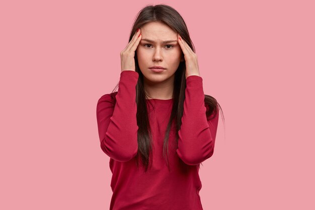 Serious beautiful young lady holds temples, feels pressured while solves puzzle in mind, wears red sweater, models against pink background, being overworked