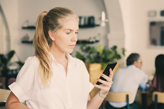 Serious beautiful young woman wearing white shirt, using smartphone, typing message, standing in co-working space
