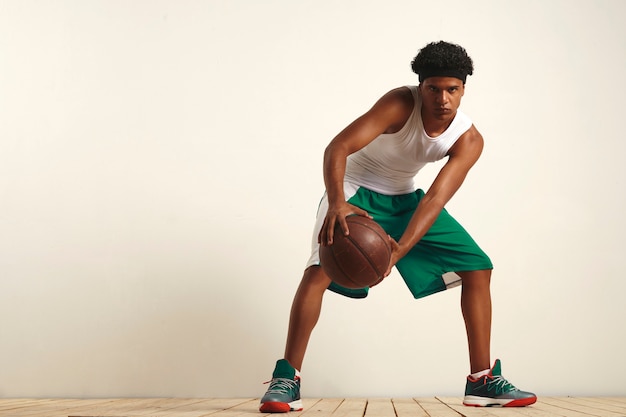 Серьезный черный спортсмен в зеленом и белом со старинным баскетбольным мячом на коленях