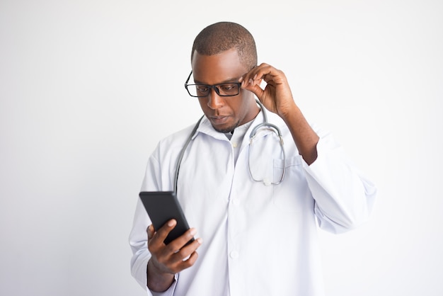 Serious black male doctor reading news on smartphone. 