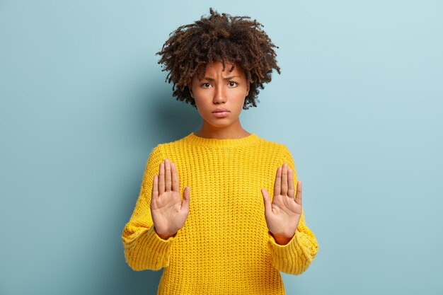 Serious black woman with grumpy expression keeps palms in front, makes stop gesture, refuses something, looks in dissatisfaction