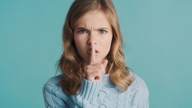 Serious blond teenage girl looking angry asking to be quiet isolated on blue background. Silence gesture