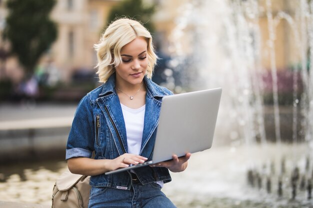 深刻なブロンドの女の子の女性学生は、日に市内の噴水の近くの彼女のラップトップコンピューターで動作します