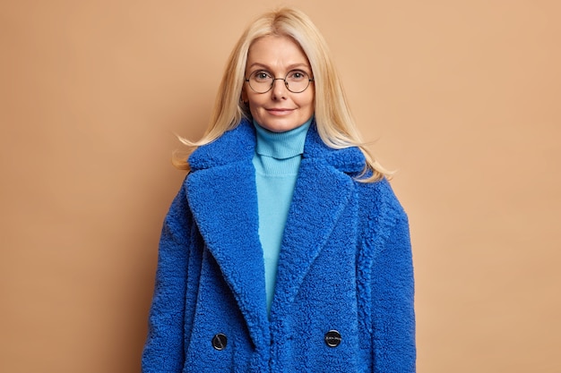 Серьезная блондинка в синей шубе с очаровательным выражением лица выглядит прямо довольной после похода по магазинам и покупки модной зимней верхней одежды.