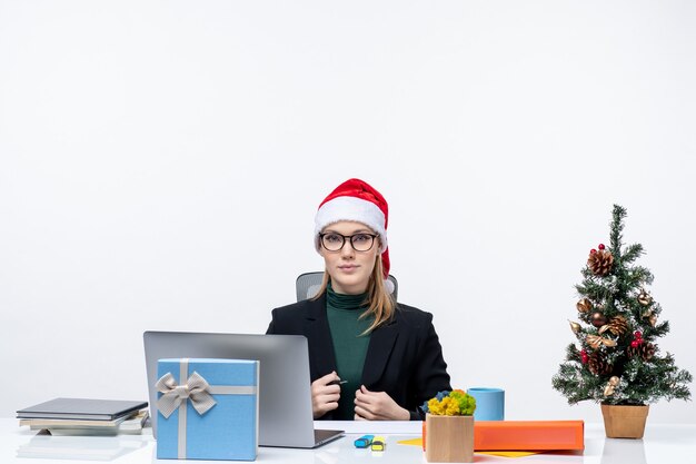 Серьезная блондинка в шляпе санта-клауса сидит за столом с елкой и подарком на ней в офисе на белом фоне