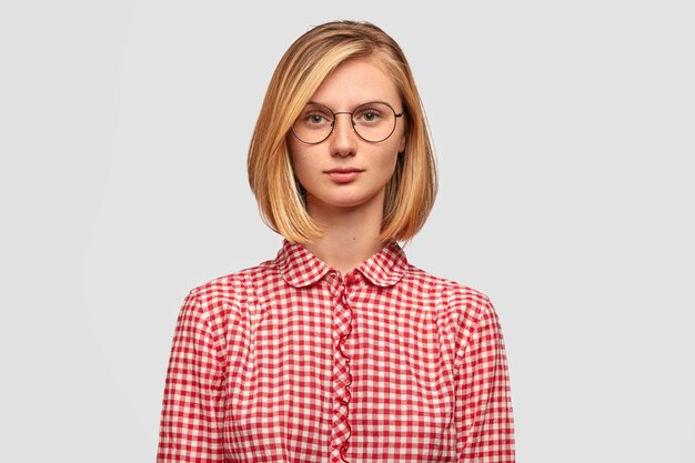 serious blonde woman with trendy hairstyle, looks seriously at camera, dressed in fashioble red checkered blouse