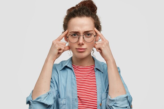 Serious blue eyed beautiful young female in spectacles, holds fingers on temples, has thoughtful clever expression, tries to remember something in mind, has freckled skin and specific appearance