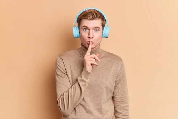 Serious blue eyed young man makes shush gesture keeps index finger over lips demonstrates silence sign asks to be quiet listens music in headphones wears casual turtleneck 