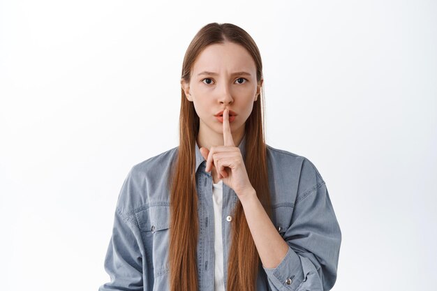 Serious bossy girl tell to be quiet, asking for silence, make shush, hush shh gesture, standing over white background. Copy space