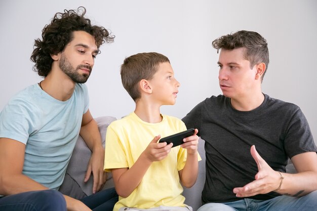 Serious boy talking to his two dads while playing game on mobile phone. Front view. Family at home and communication concept