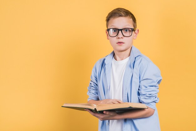 黄色の背景に対してカメラを探して手に開いた本を持って眼鏡をかけている深刻な少年