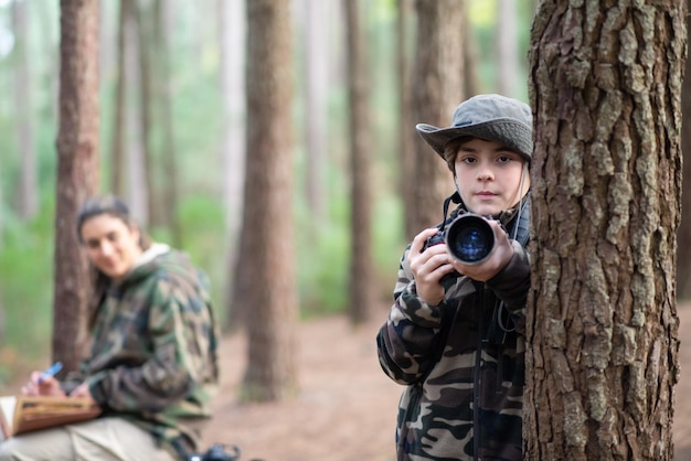 無料写真 森の中でカメラを持つ深刻な少年。カメラを見ているコートとパナマの男子生徒。バックグラウンドでぼやけた母親。子供の頃、自然、レジャーの概念