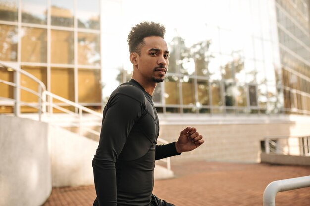 Serious brunet curly darkskinned man in black sport longsleeved shirt looks into camera and runs outside