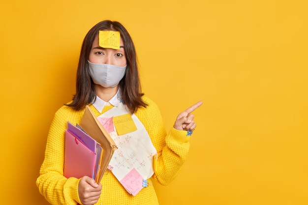 Серьезная брюнетка Азиатская женщина с наклейкой на голове пытается узнать информацию для сдачи экзамена, успешно носит одноразовую маску, чтобы предотвратить коронавирус, указывает на пустое пространство