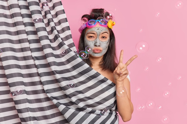 Serious brunette Asian woman makes peace gesture hides naked body behind shower curtain poses in douche applies clay mask for skin refreshment