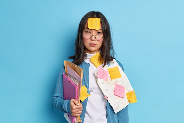 Serious brunette Asian woman with sticker stuck on forehead busy doing paperwork prepares financial report wears round spectacles casual jumper has clever look.