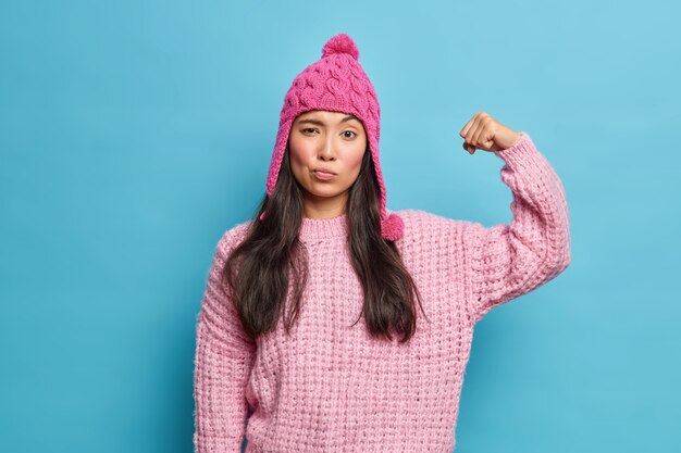 Serious brunette woman raises arm and shows muscles being self confident and full of power wears knitted jumper pink hat feels strong healthy isolated over blue wall