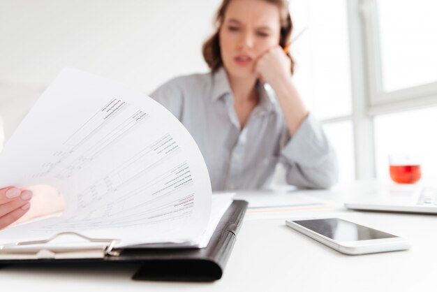 serious brunette woman reading papers while siting at workplace in light aparment, selective focus on document