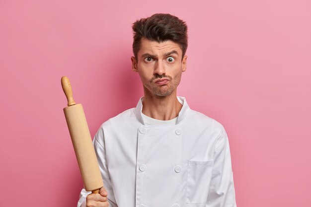 Serious brutal man chef wears white uniform holds wooden rolling pin