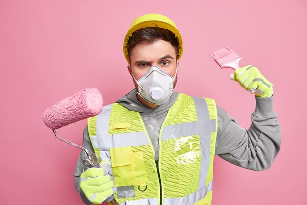 Serious builder worker holds paint roller and brush wears hardhat protective mask uniform works on repairing of new house poses against pink wall. Building renovation and reconstruction