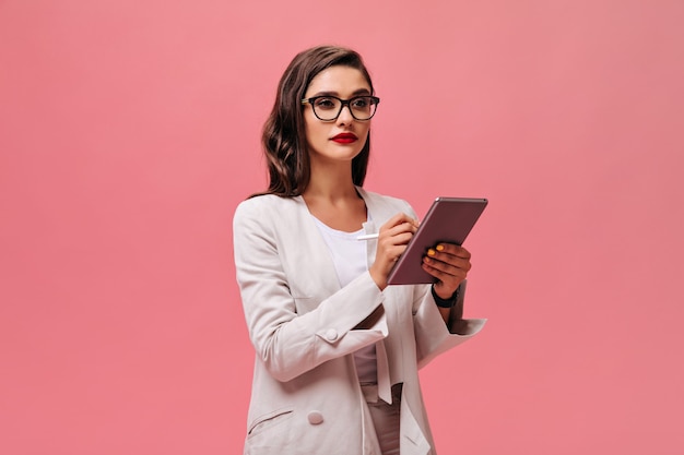 Серьезная бизнес-леди с ярко-красными губами в бежевом стильном наряде и очках держит планшет на розовом изолированном фоне.