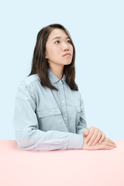 Serious business woman sitting at table, looking up isolated on trendy blue studio background. Female half-length portrait.