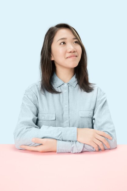 Serious business woman sitting at table, looking up isolated on trendy blue studio background. Female half-length portrait.