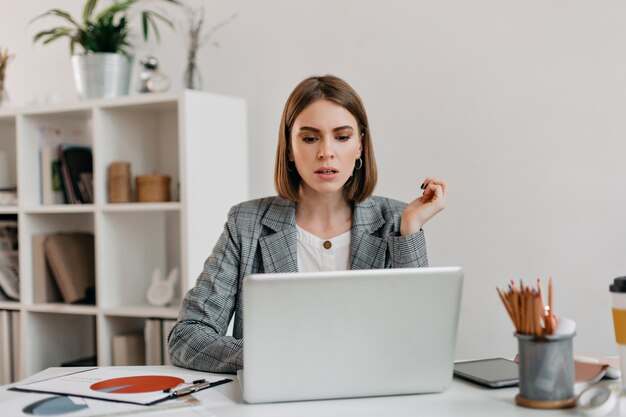 Serious business woman with anxiety looks in laptop. Portrait of girl with short haircut in white office.