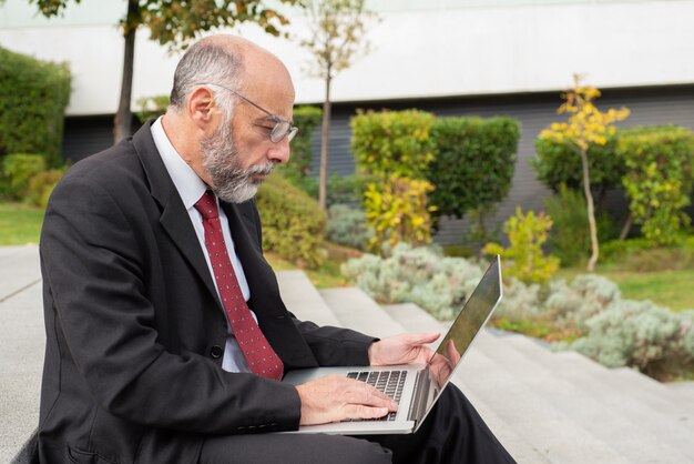 Serious businessman in eyeglasses using computer on street