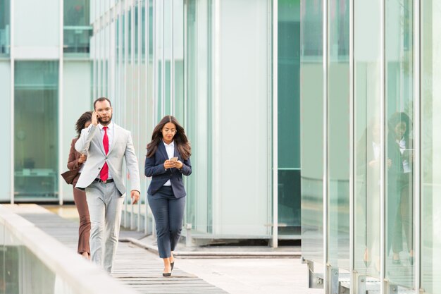 Serious businesspeople with gadgets going along urban glass wall