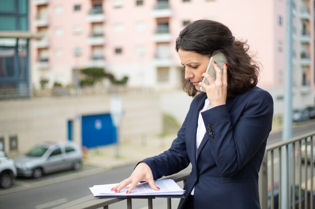 Серьезная деловая женщина разговаривает по смартфону и смотрит на документы