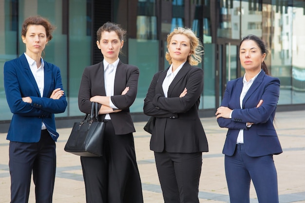 Serious businesswomen group with arms folded standing together near office building, posing, looking at camera. Front view. Business team or teamwork concept