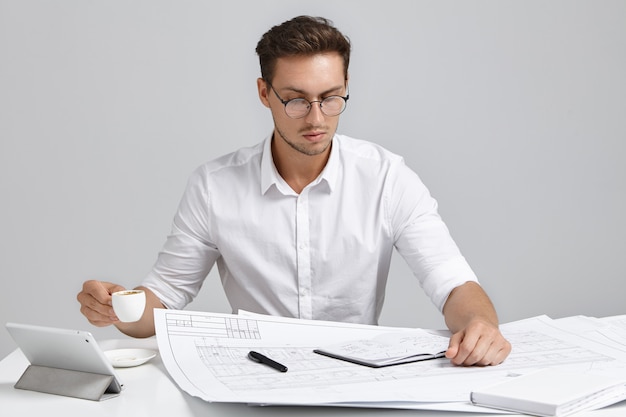 Serious busy male designer dressed formally, looks attentively into blueprints, drinks coffee or espresso.