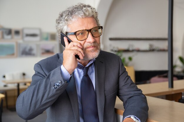 Serious busy mature businessman wearing glasses, talking on mobile phone, standing at co-working, leaning on desk, looking away