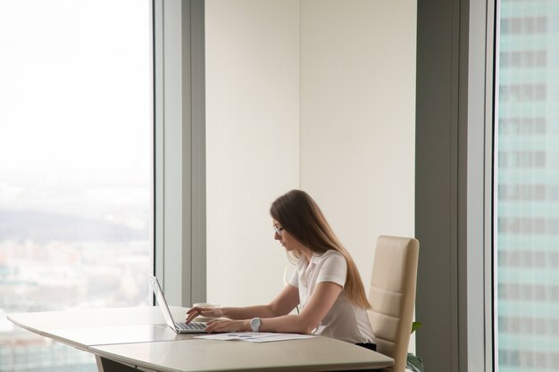 Serious busy woman working on laptop in office interior, copyspace