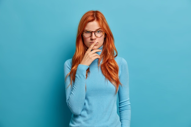 serious calm woman has natural long ginger hair keeps hands on lips and looks with concentrated thoughtful expression.