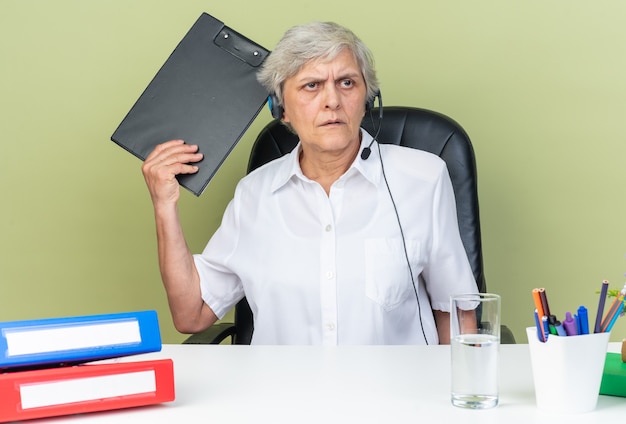 Serious caucasian female call center operator on headphones sitting at desk with office tools holding clipboard looking at side isolated on green wall