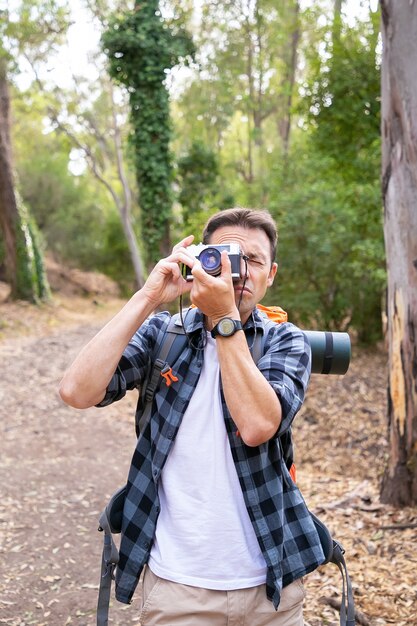 Serious Caucasian man shooting landscape and hiking in forest. Male traveler walking on nature, taking photo and standing on road. Tourism, adventure and summer vacation concept