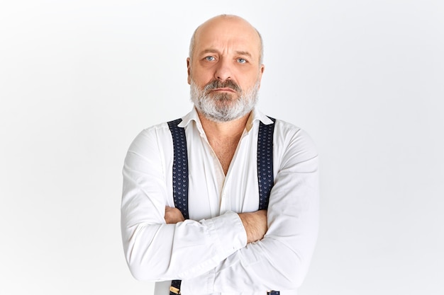 Serious Caucasian retired businessman wearing white shirt and suspenders looking at camera with confident facial expression, crossing arms on his chest