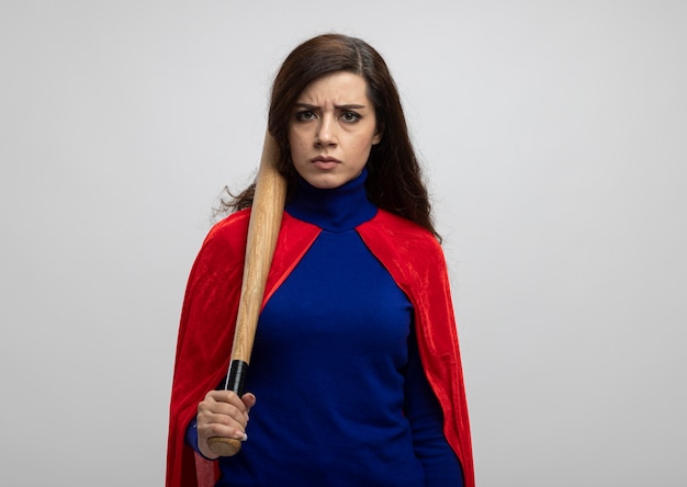 Serious caucasian superhero girl with red cape holds baseball bat isolated on white wall with copy space