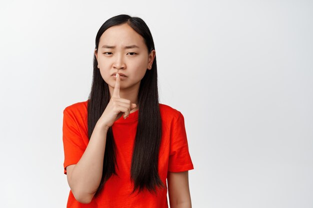 Serious chinese girl looking angry shh at camera press finger to lips and shushing tell to be quiet scolding person standing over white background