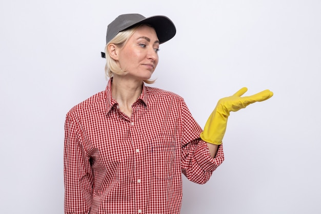 何かを提示している彼女の腕を見ているゴム手袋を着用している格子縞のシャツとキャップの深刻な掃除の女性