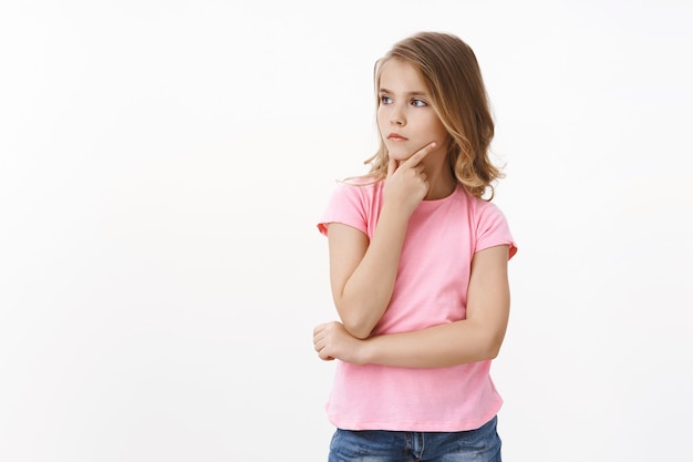 Serious clever lovely little girl with blond hair, daughter thinking what draw, search inspiration, touch chin thoughtful, ponder decision, look left intrigued, pick what buy, stand white wall