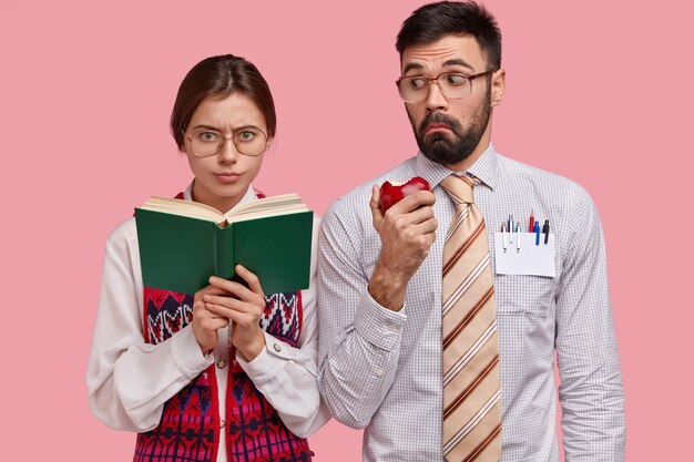 Serious clever schoolgirl in thick lenses spectacles holds handbook, studies indoor, surprised bearded man in formal shirt