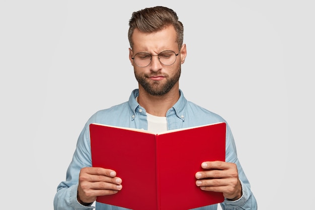 Serious clever young male teacher with trendy haircut, carries red book, loos hesitant