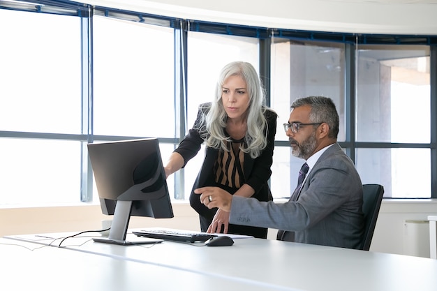 Serious colleagues watching and discussing content on computer monitor, pointing at display and talking while sitting in meeting room with panoramic window. Business communication concept