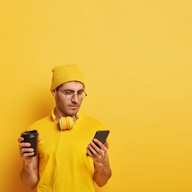Серьезный сосредоточенный парень держит мобильный телефон, читает новости в интернете, одет в желтую шляпу и футболку, пьет ароматный напиток, подключен к бесплатному Wi-Fi