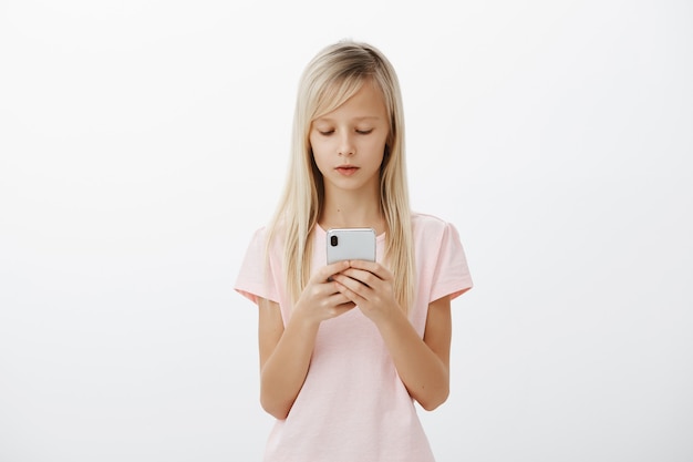 大人っぽく振る舞うまじめな女の子。ブロンドの髪、スマートフォンを押しながら画面を見たり、漫画を見たり、灰色の壁越しにメッセージを送ったりして焦点を合わせた愛らしい子供の屋内撮影